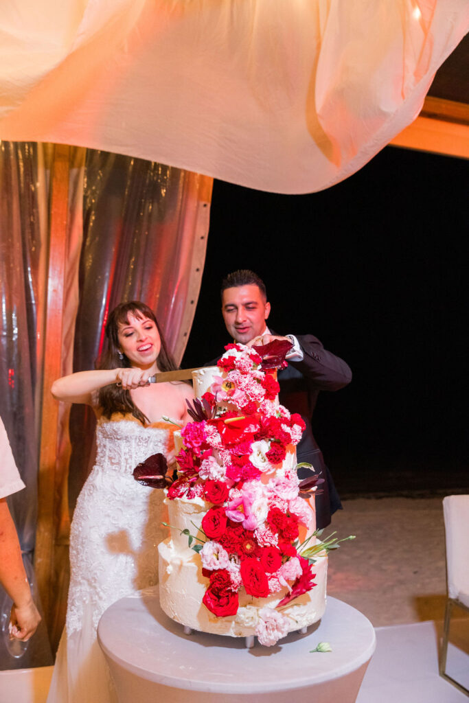 Bride and groom cutting a tall floral wedding cake with red and pink roses during their destination wedding reception.