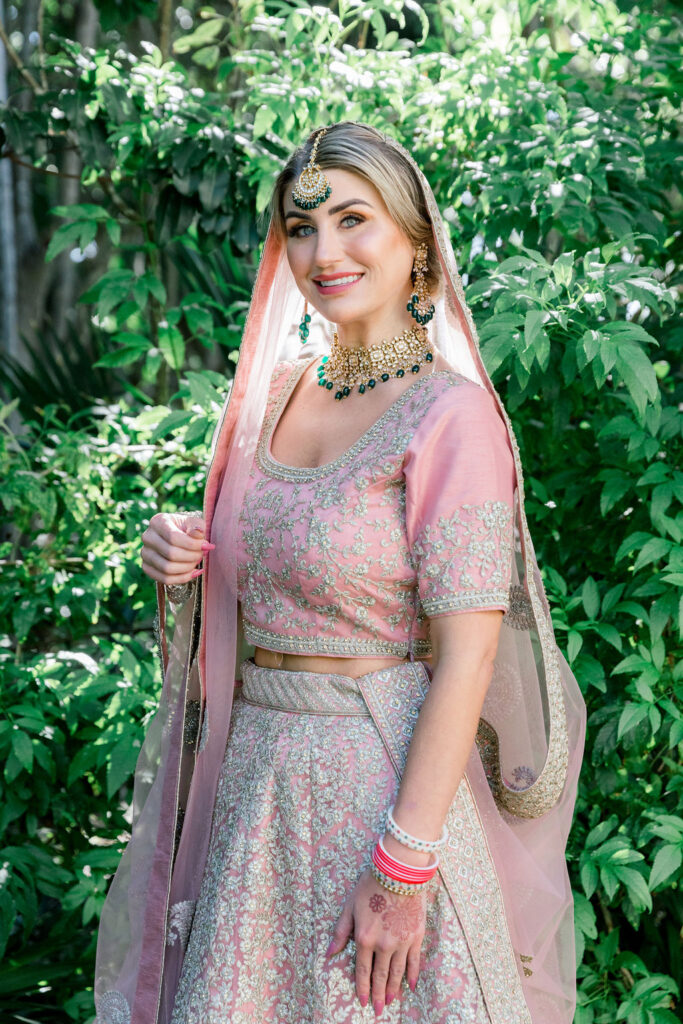 Destination wedding bride wearing a traditional pink and silver embroidered lehenga with jewelry and veil, photographed outdoors by an international wedding photographer.
