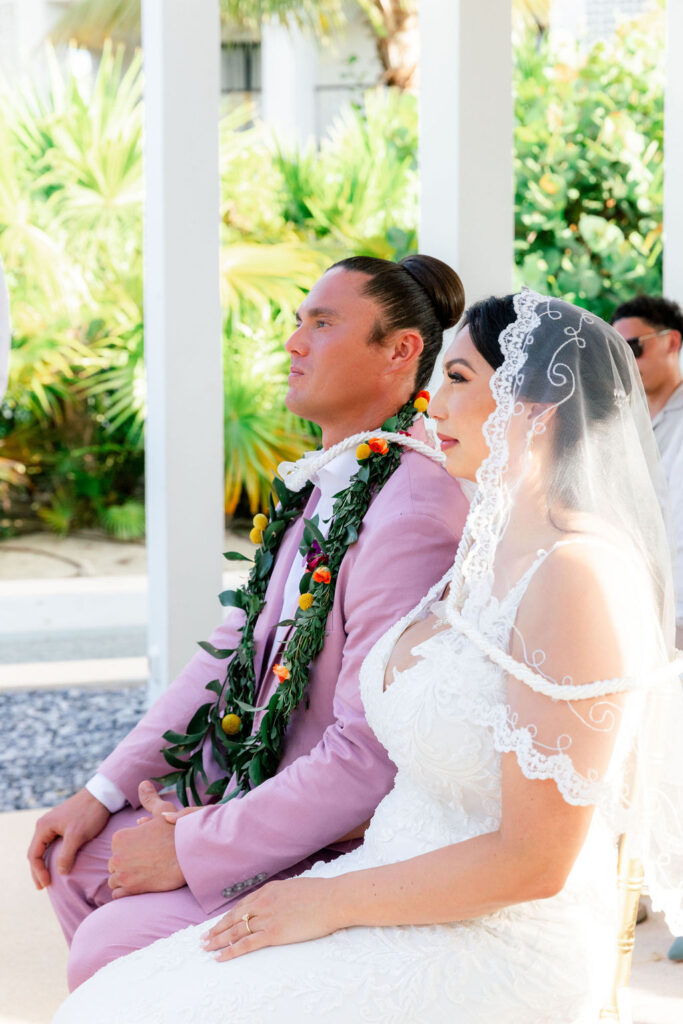 Multicultural wedding photography of a bride in lace gown with veil and groom in pink suit wearing floral garlands during ceremony in Riviera Maya.