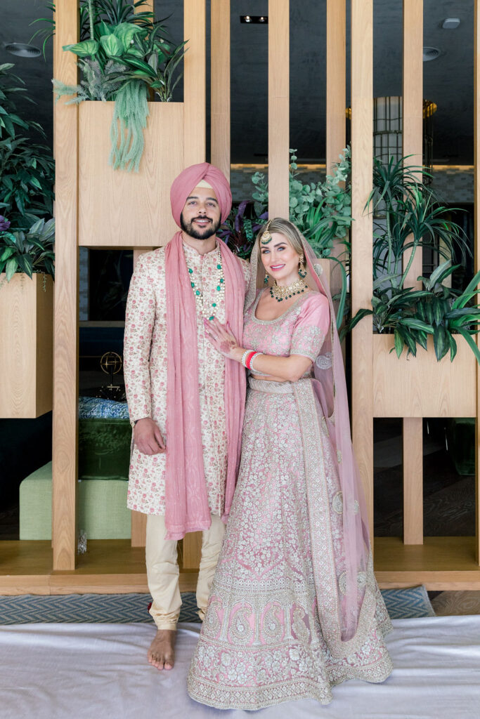 Couple in traditional Indian wedding attire, groom wearing sherwani and turban with pink scarf and bride in embroidered lehenga with matching jewelry, photographed at a multicultural destination wedding.