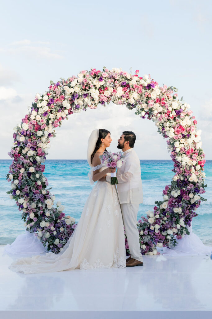 Bride and groom at a beachside multicultural wedding in front of a floral circular arch with purple, pink, and white flowers, captured by a destination wedding photographer in Mexico.