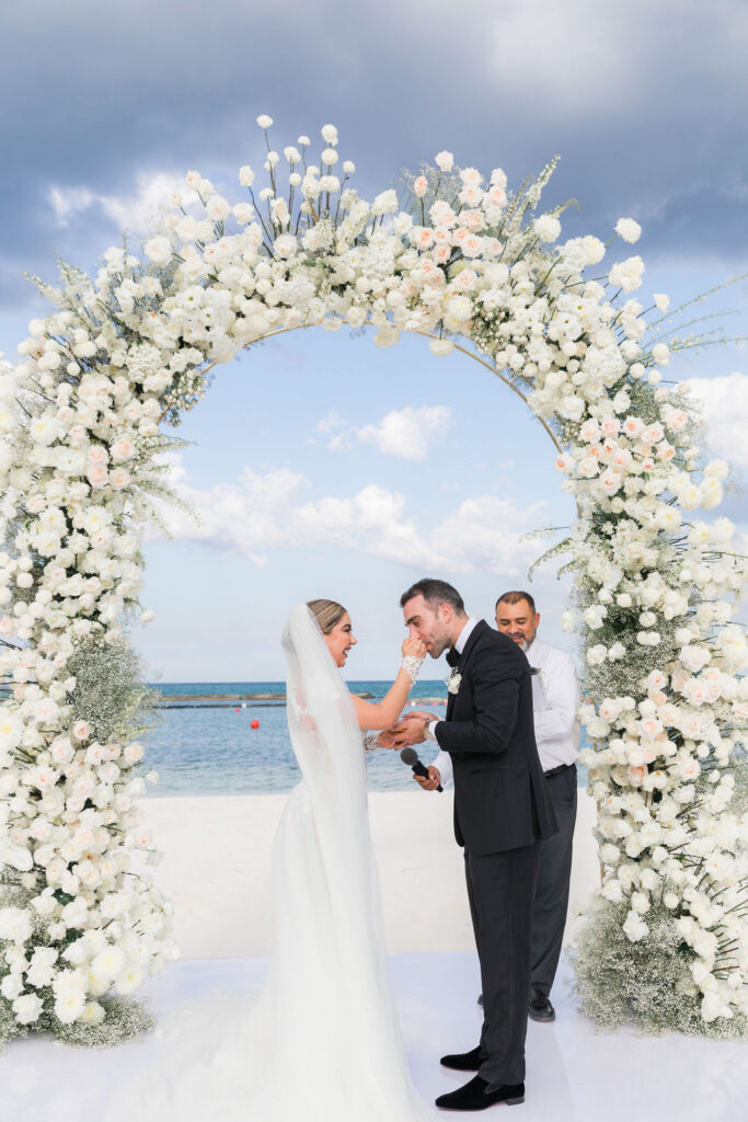 Multicultural destination wedding ceremony by the sea, bride in a white gown with veil and groom in black suit under a circular arch of white roses, photographed by a luxury wedding photographer.