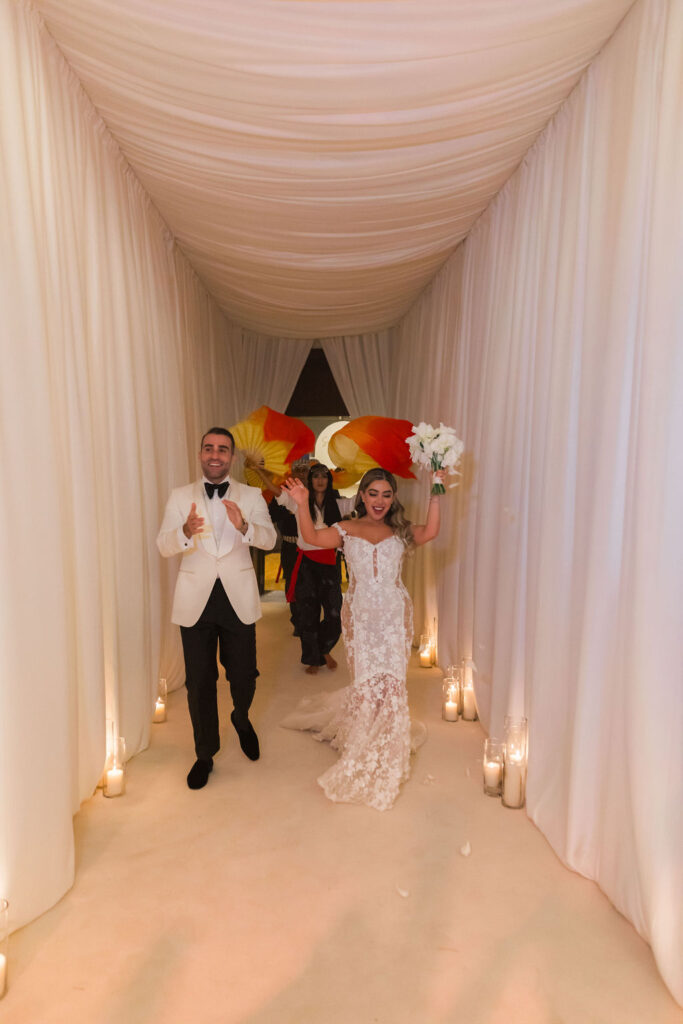 Bride and groom entering their multicultural destination wedding reception through a candlelit white draped hallway, captured by a luxury wedding photographer.