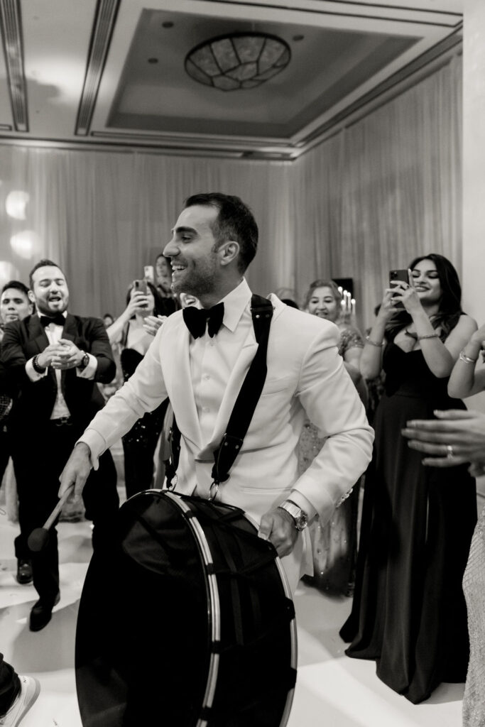 Black and white photo of groom playing a drum surrounded by guests during multicultural wedding celebration, documented by a destination wedding photographer.