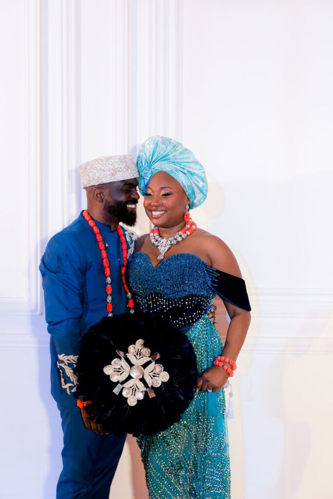 Multicultural destination wedding couple in traditional African attire, bride in turquoise gown with headwrap and groom in blue outfit with coral beads, captured by a luxury wedding photographer.