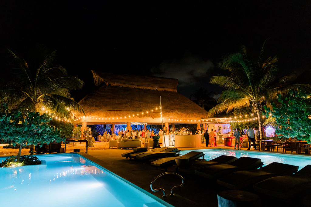 Outdoor wedding reception by the pool at night, illuminated with warm string lights under a tropical palapa roof.