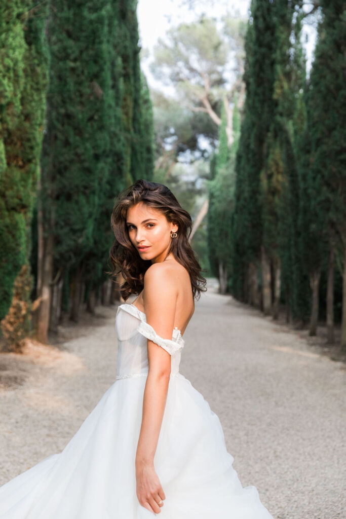 Bride in a voluminous white gown walking through a cypress-lined path, photographed in soft natural light for an editorial-style wedding portrait.
