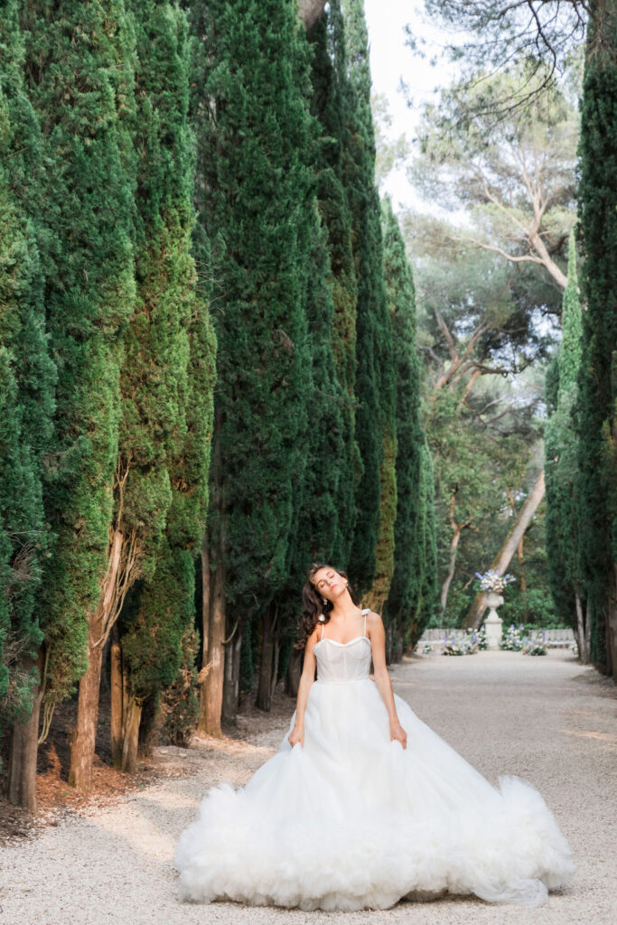 Fashion-inspired bridal portrait of a bride in an off-the-shoulder gown standing between tall cypress trees, captured in warm natural light.