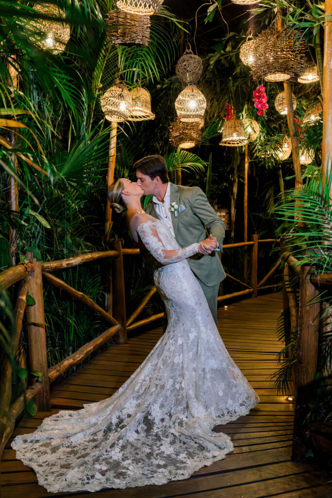 Bride and groom dancing on a wooden bridge surrounded by lush tropical plants and hanging lanterns, photographed in warm nighttime lighting.